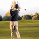 A golfer in a navy sweater and beige pants swings a club on an open golf course. The background features scattered trees and a cloudy sky. A golfer in a navy sweater and beige pants swings a club on an open golf course. The background features scattered trees and a cloudy sky.